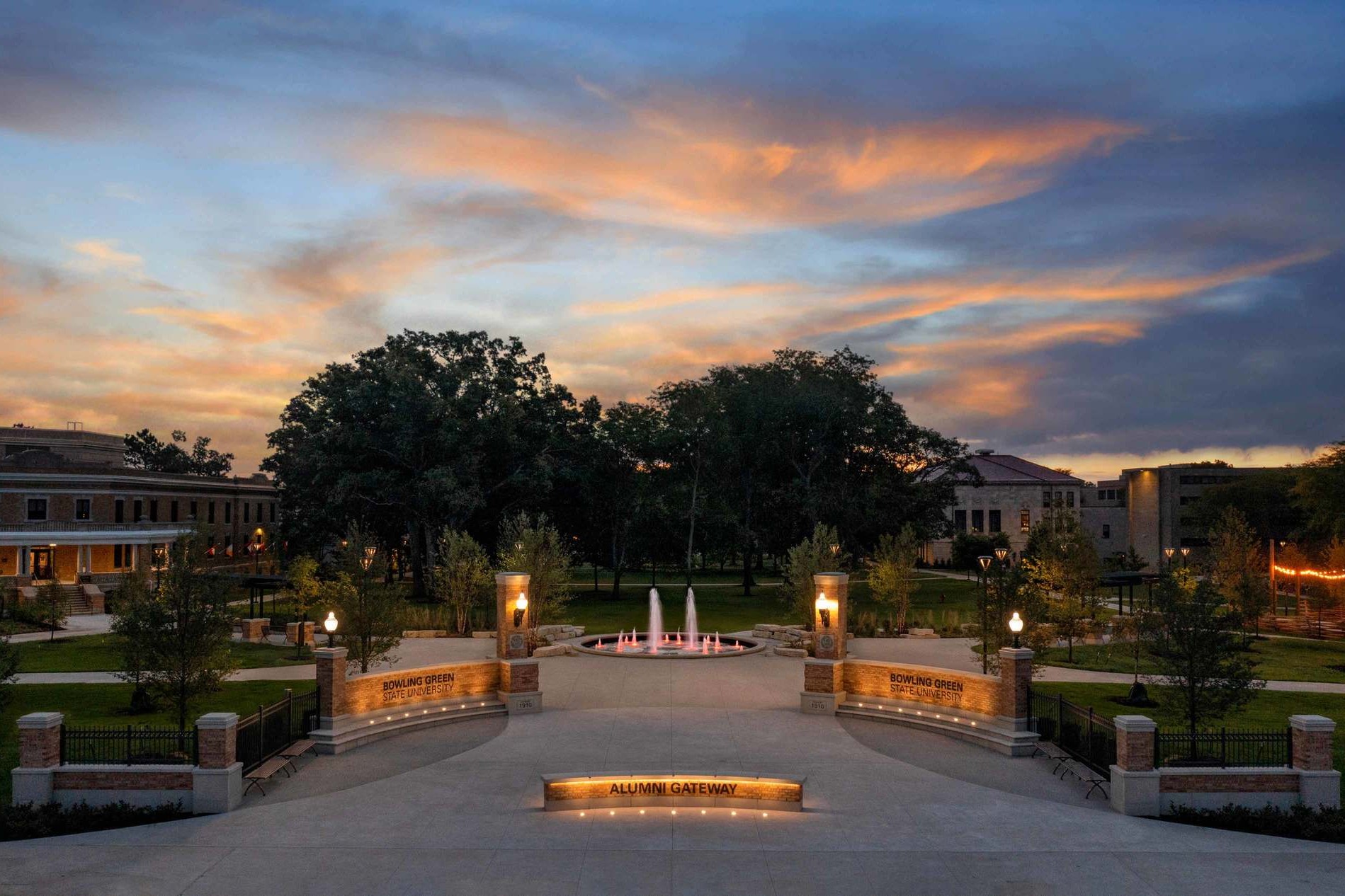 A photo of Bowling Green State University campus at night, an orange and blue sunset in the sky. At center, a fountain surrounded by brick campus buildings.