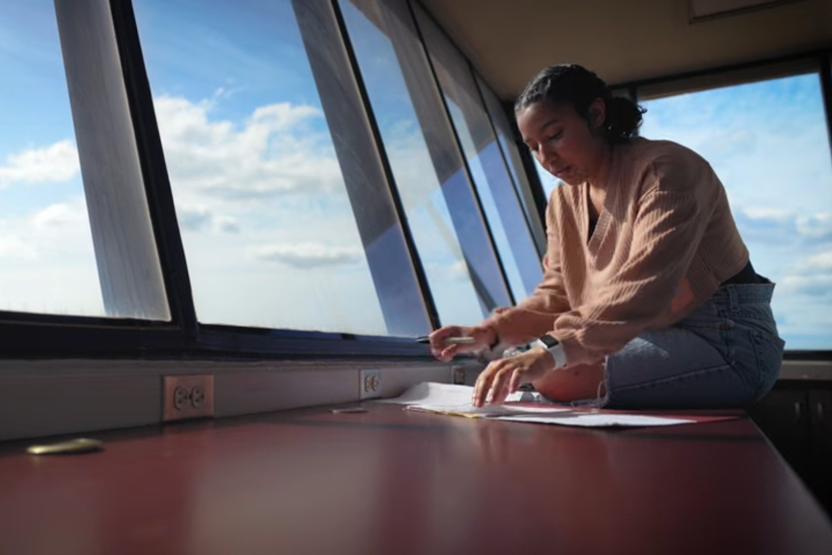 Aria Vargyas sits on a counter in a weather station sorting papers. The windows beside her show blue skies.