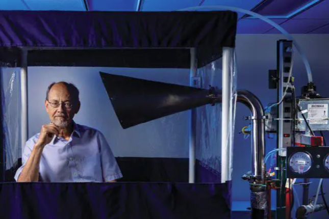 Donald Milton sits inside a chamber constructed of clear plastic tarp and blue cloth. A funnel inside the chamber is connected to scientific equipment sitting outside it.
