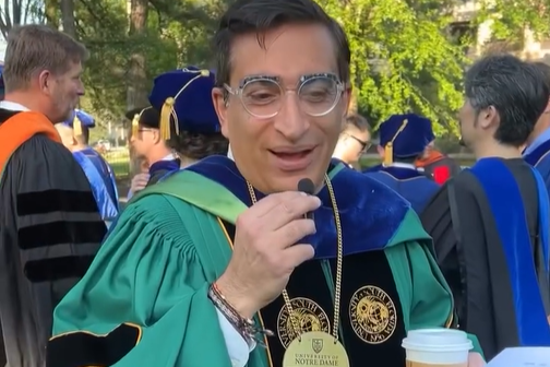 A professor dressed for commencement in green robes holds a tiny mic and answers a question for the camera.