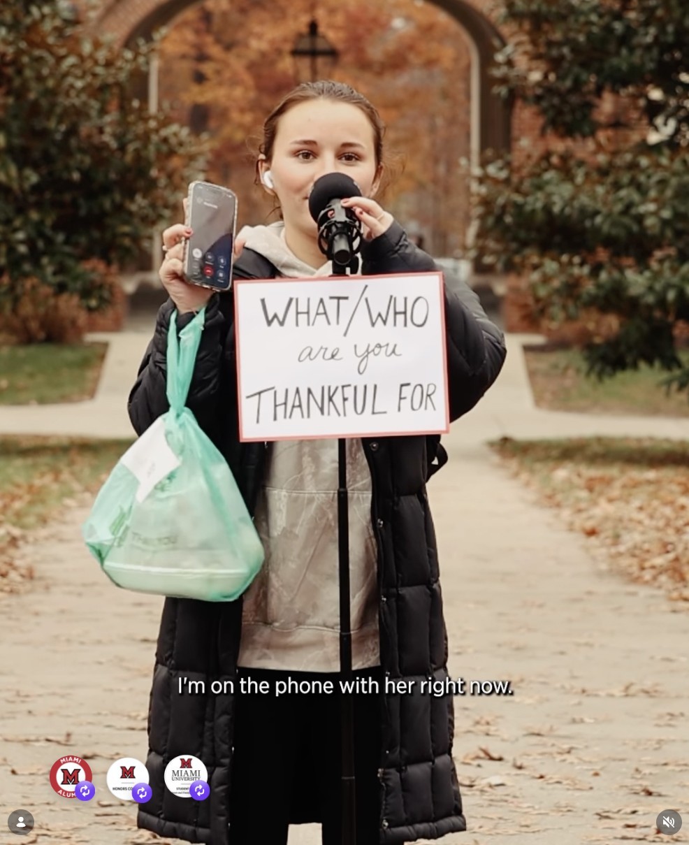 A screenshot from Miami University's "Who or What Are You Thankful For?" video shows a girl in a winter coat holding her phone up to camera as she speaks into a microphone, saying, "I'm on the phone with her right now."