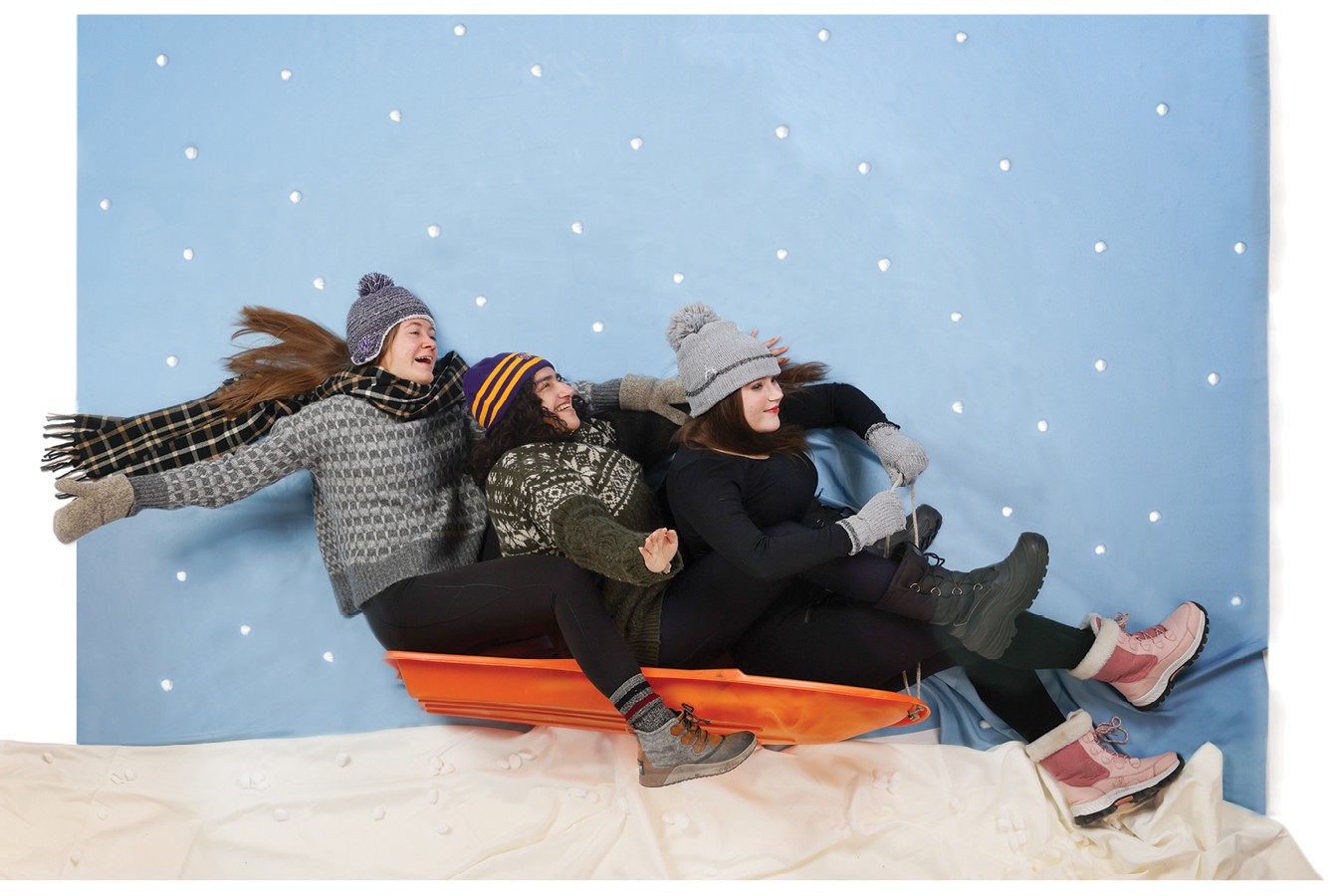 Three girls sit on a sled wearing winter hats, sweaters, and snow boots. They are posed to look like they are in motion sledding down a hill.