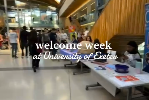 A screenshot from a video of the interior of a campus building with tables set up along its edges. The text, "welcome week at University of Exeter" has been placed at the center of the screen.