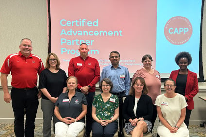 A cohort poses for a photo in two rows in front of a presentation screen that reads "Certified Advancement Partner Program."
