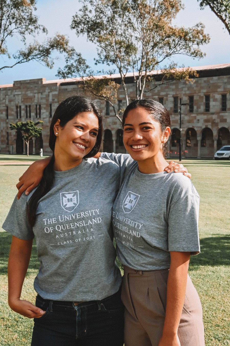 Two UQ alumna pose with their arms around each other while wearing unique gray with white embroidery class shirts.