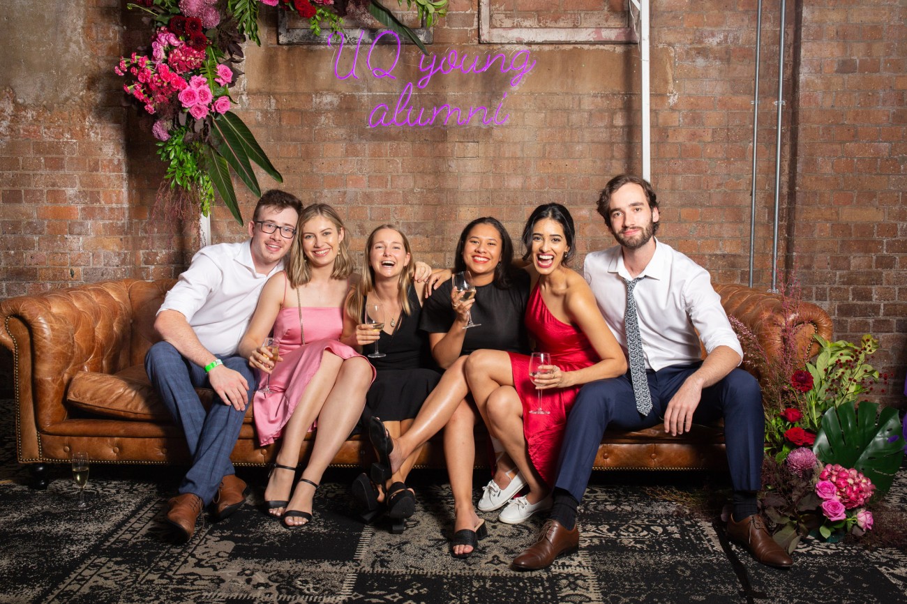 A group of UQ alumni and friends pose on a brown leather couch to have their photo taken at the UQ 2021 Party with a Purpose event. Above them hangs a purple neon cursive font sign that reads "UQ young alumni."
