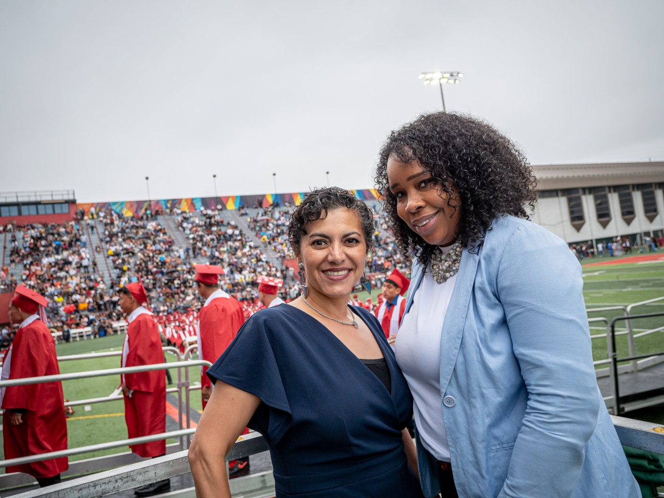 Two women pose together on a football field, a commencement ceremony taking place in the background.
