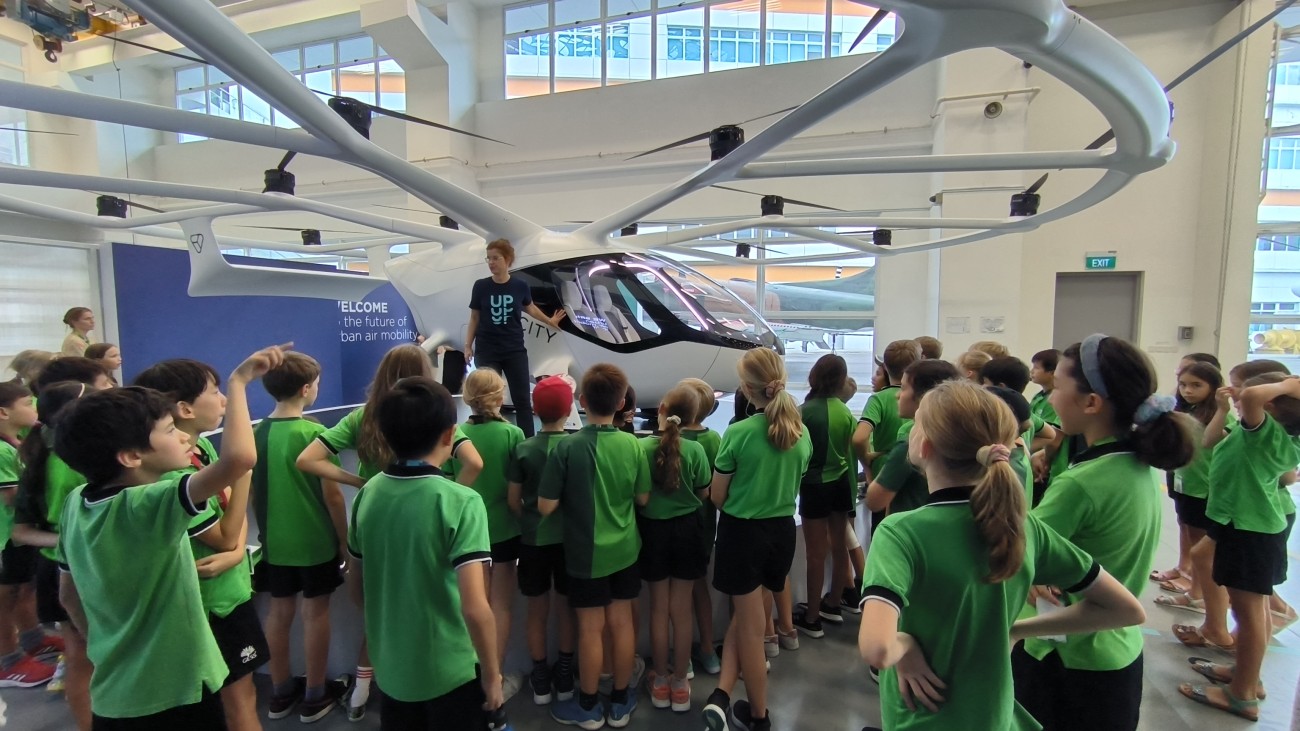 A group of students wearing green matching green shirts crowd around a platform to view a Volocopter drone.