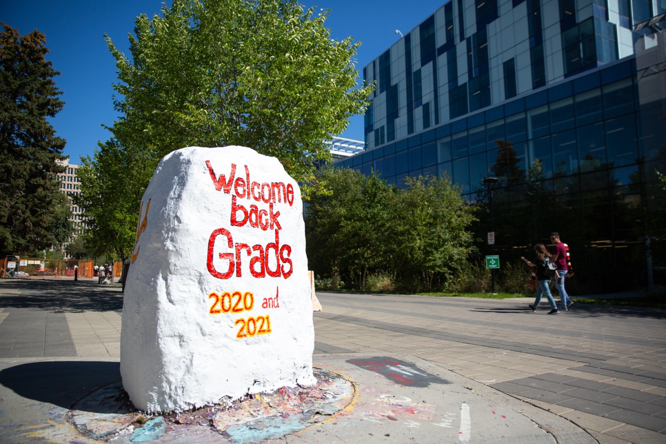 A rock painted white with the words "welcome back grads 2020 and 2021" on University of Calgary's campus