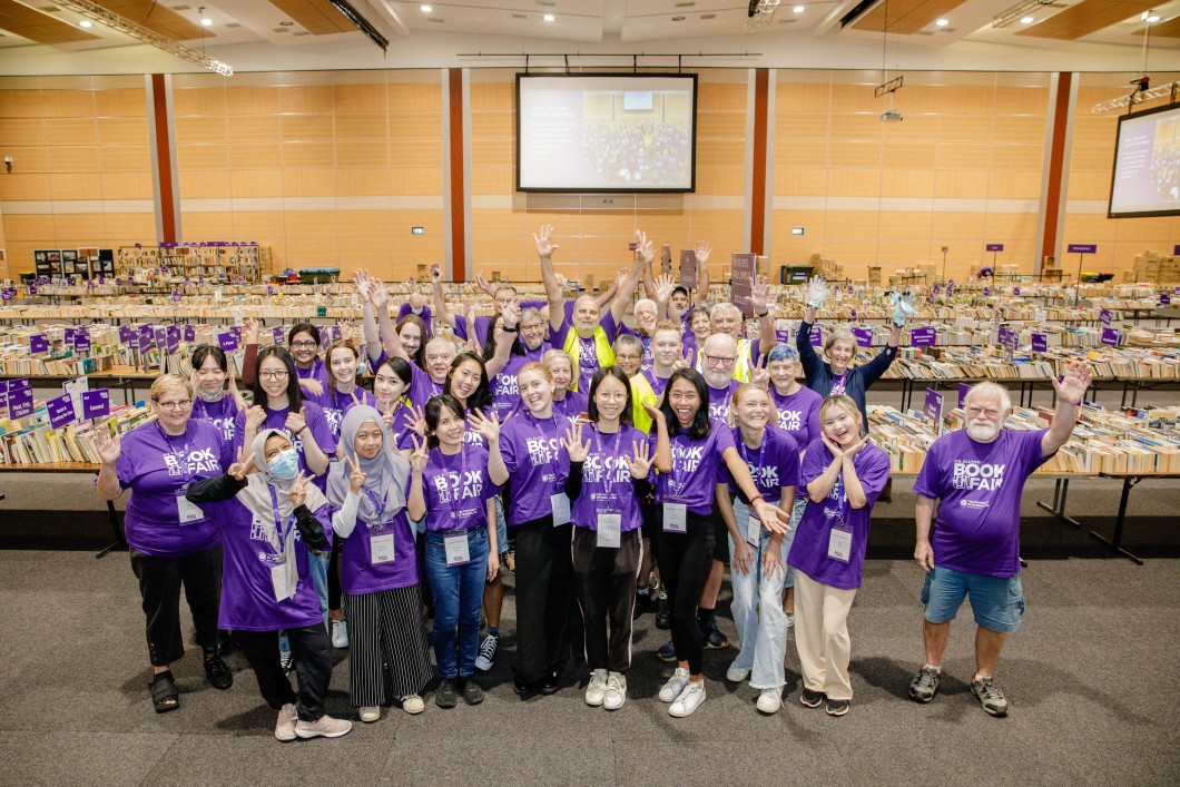 UQ Alumni Book Fair volunteers, all wearing matching purple shirts, pose together for a fun group photo on the book fair floor.