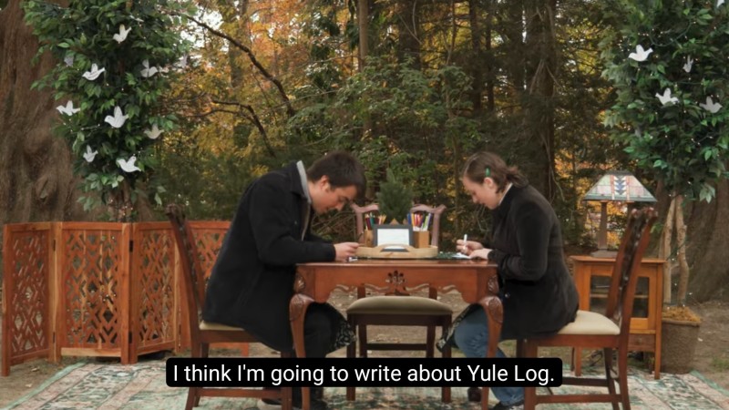 Two William & Mary students sit at an ornate table writing cards. One of them says, "I think I'm going to write about Yule Log."
