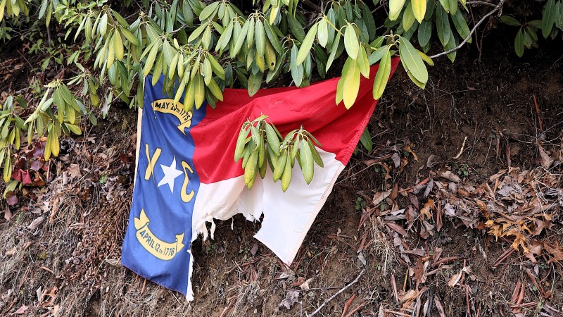 A North Carolina flag with its bottom right corner torn off hangs from branches against a muddy hillside.