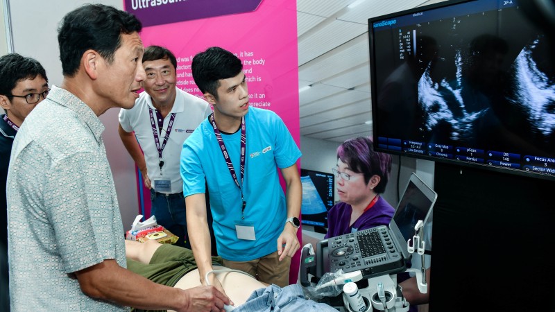 Four people gather around a table to perform an ultrasound on a fifth person. A medical professional sits next to the ultrasound machine.