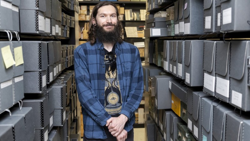 A photo of Tyler Lee-Wynant, a UC Berkeley PhD student, standing between two tall shelves full of collection material.