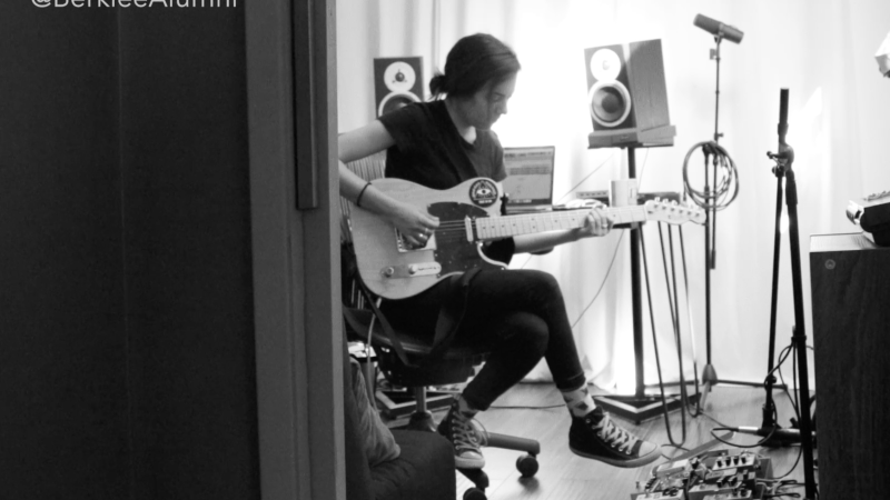 A black and white photo of a student playing guitar with studio equipment surrounding them.