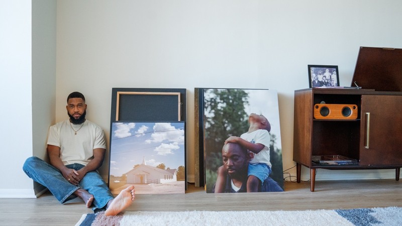 John Johnson sits on the floor next to prints of his own photography. Further along the wall is a wooden record stand and player.