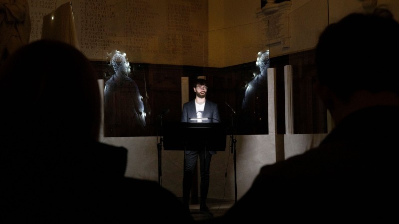 A man performs a reading in a darkened room, images of Byron projected behind him.