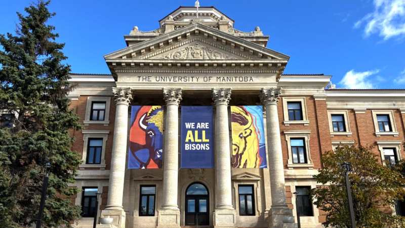 A photo of a columned building on the University of Manitoba campus. A series of three red, blue, and yellow banners hang between the building's columns. The right and left banner shows images of a bison, with the middle banner reading "We are all Bisons."
