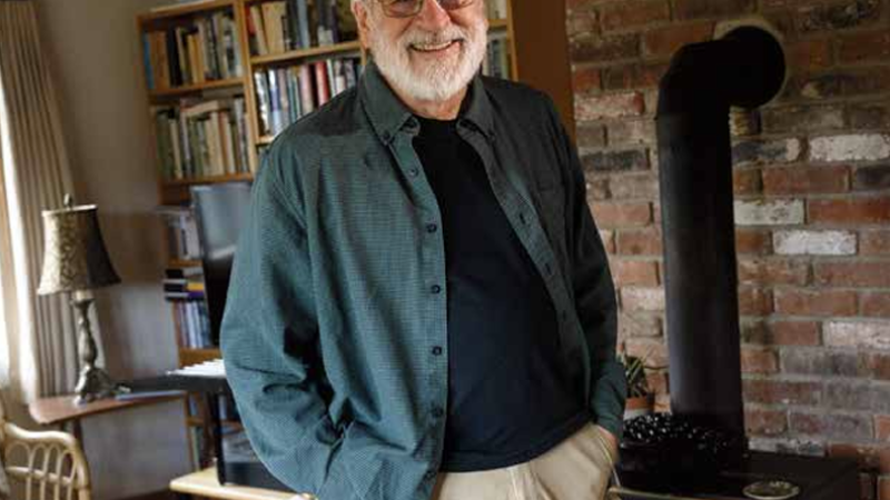 A photo of Tom Lee in his house, hands in his pockets. Behind him is an antique stove and bookshelves.