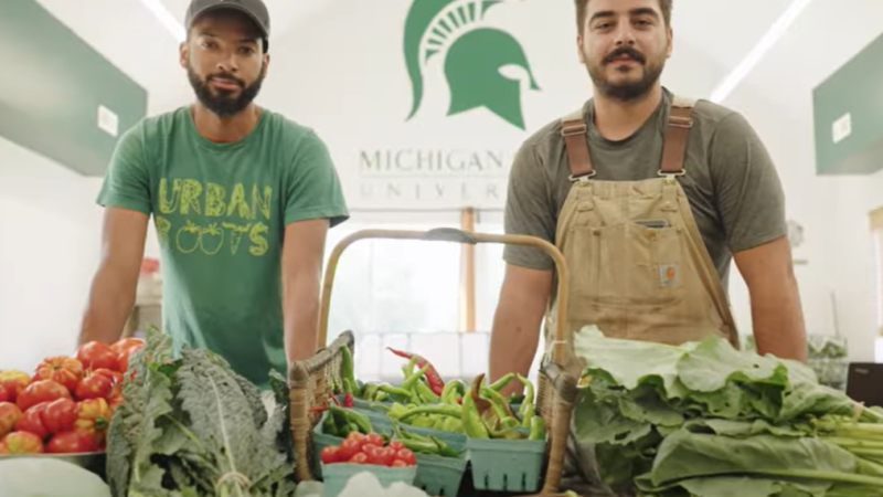 Two people wearing Michigan-green stand behind a table covered in fresh vegetables, including tomatoes, peppers, and leafy greens.