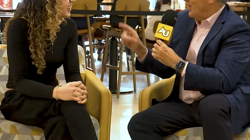 In a coffee shop, Board Chair Michael A. L. Balboni and Adelphi student Emily Munayirji sit in side by side chairs. Michael holds a microphone, interviewing her.
