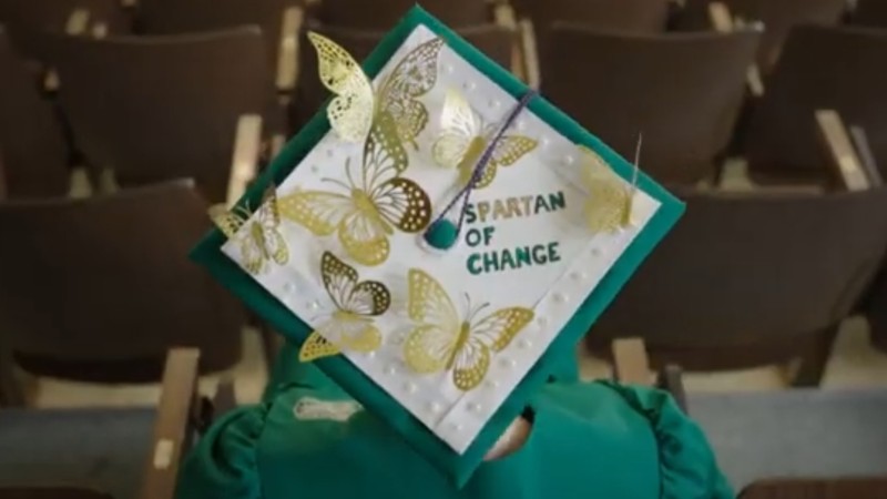 The top of a Michigan State graduation cap is decorated with gold foil butterflies and the words "Spartan of Change" in green foil.