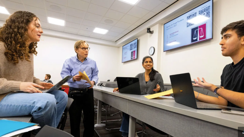A photo of three students with laptops and a professor conferring in a classroom.
