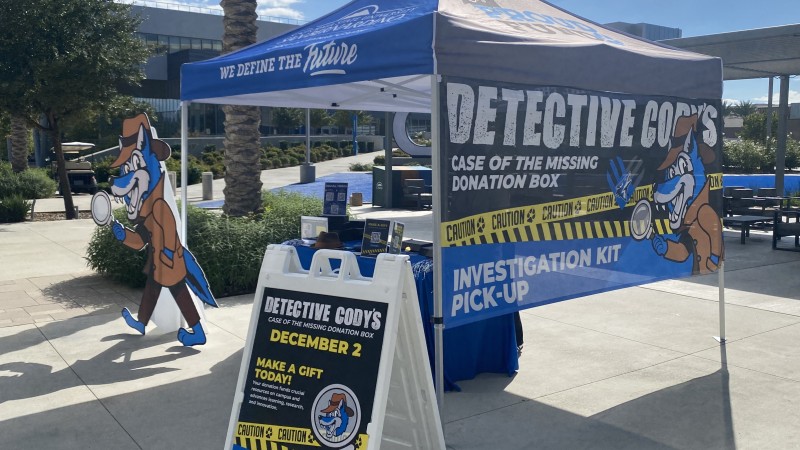 A photo of a blue tent set up for CSUSB Giving Day. Next to the tent is a cardboard cut out of the Detective Cody character, a blue wolf wearing a trench coat and a deerstalker hat and holding a magnifying glass.