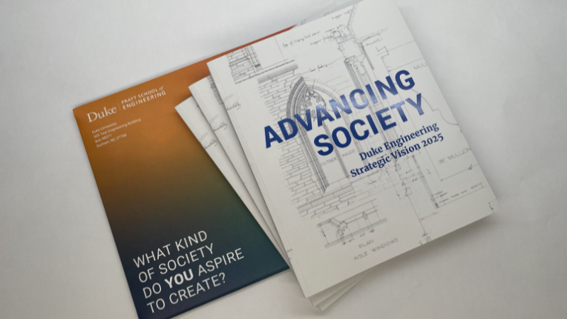 A photo of the Duke Engineering Strategic Vision publication and the orange and blue ombre colored envelope it was sent in. The cover of the publication features blue-colored text that reads, "Advancing Society," and a white background with black architectural drawings.