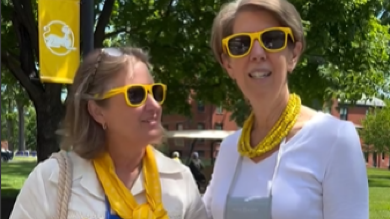 Two women wearing yellow sunglasses talk to camera. In the background, a yellow banner for MHC hangs from a light pole in the middle of a sunny and grassy campus.