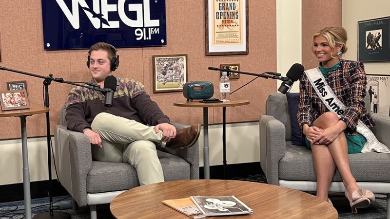 On the set of the "Everything Auburn" podcast, one of the hosts and a guest, a Miss America contestant, sit in chairs with microphones in front of them.