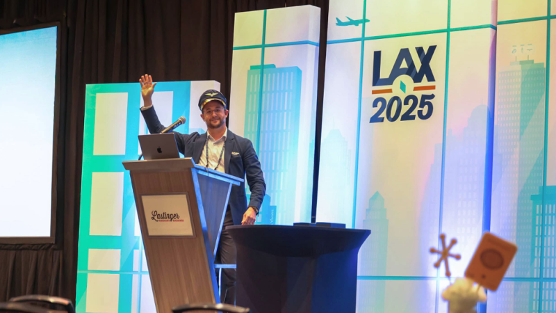 A photo of a man standing on stage at a podium wearing a sailor's hat. Behind him is a white and blue graphic with images of a skyline, a plane, and the text "LAX 2025."