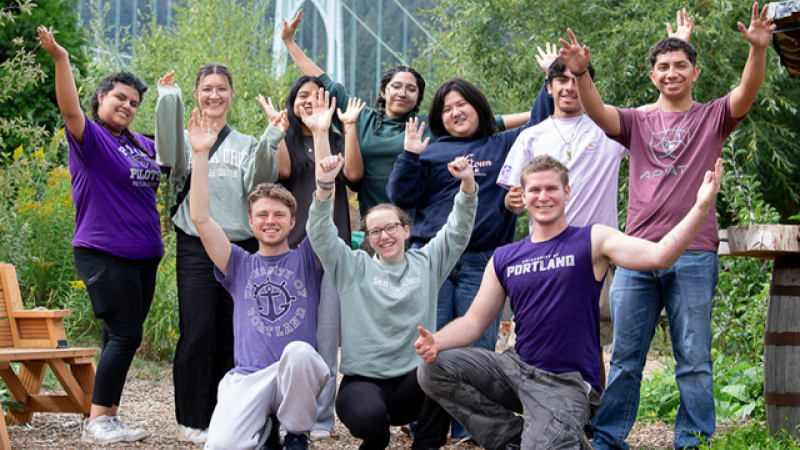 A group of University of Portland students dressed in university merch smile for camera with their hands in the air.