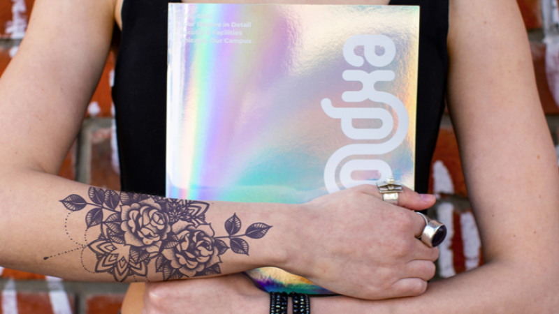 Close up of someone holding SAIC's explore booklet in front of their chest. The shiny silver cover reflects in the sun.