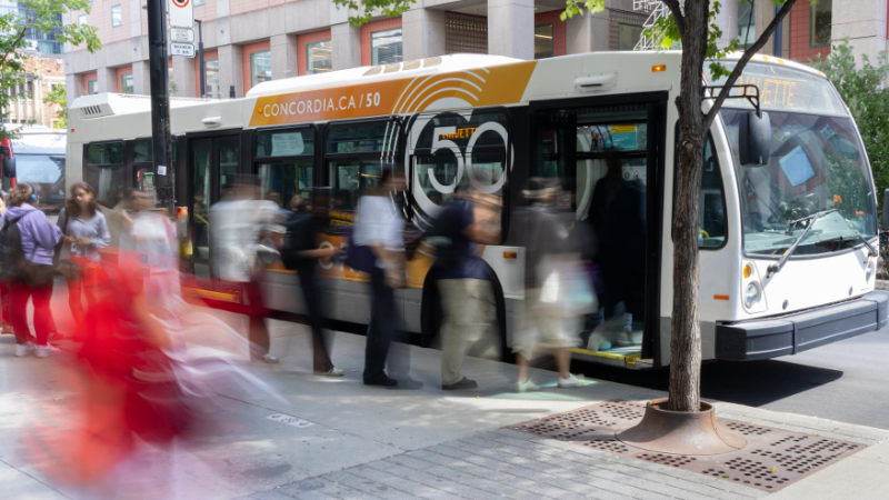 Photo of a shuttle bus with Concordia's 50th Anniversary-branded advertisement on its side. A line of people wait to enter the bus.