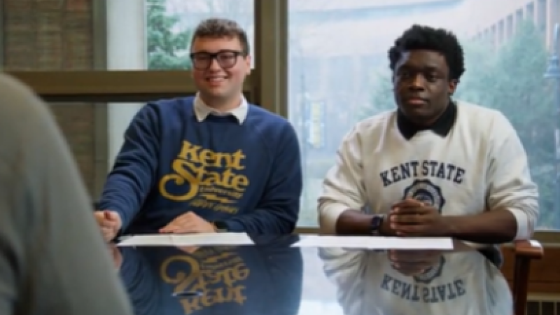 Two Kent State students wearing Kent State sweatshirts sit across the table from a campus leader as they pitch their video script.