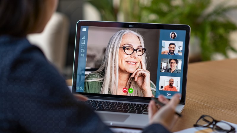 A woman is looking at a laptop screen, with a video meeting on it. She's chatting with four other participants on the screen.