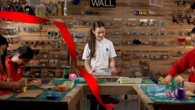 Screenshot from HKIS's promotional video. Students paint and craft at three tables shaped in a "U." A animation of a red ribbon flies through the screen.
