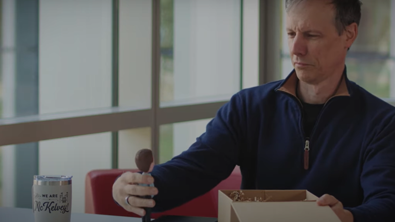 Sitting at a table, a man removes a bobble head of himself from a giftbox.