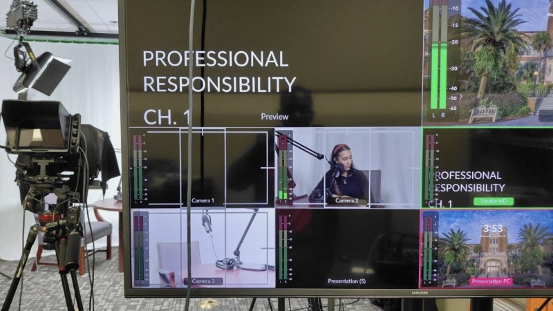 A photo shows a screen that displays multiple camera set-ups for the recording of a video. Behind the screen is a camera and chairs in front of a white back drop.