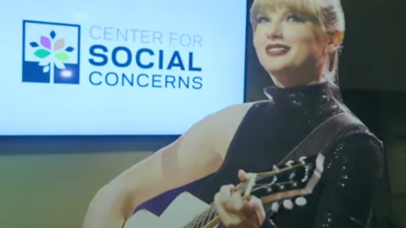 A cutout of Taylor Swift holding a guitar mid-performance is held up in front of a screen where the words "Center for Social Concerns" are displayed.