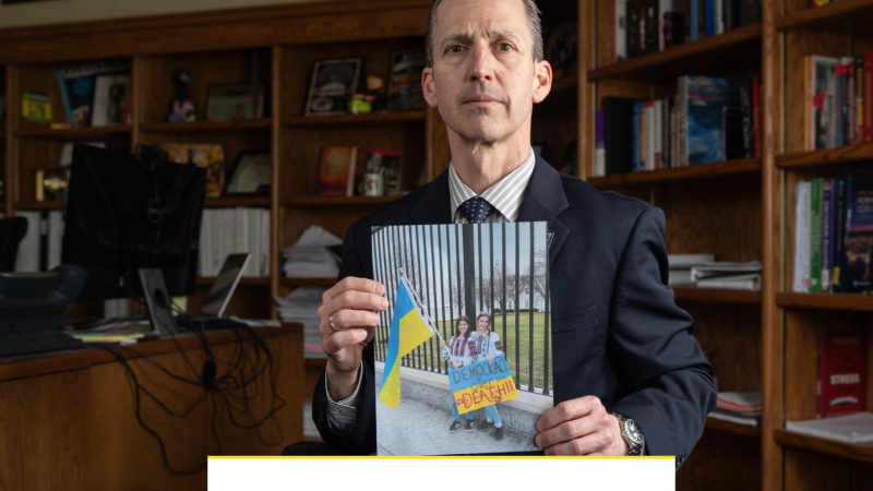 A man stands in the middle of an office holding a photo of two girls outside a fenced in property. One holds a Ukrainian flag and the other a poster that reads "Democracy over Death."