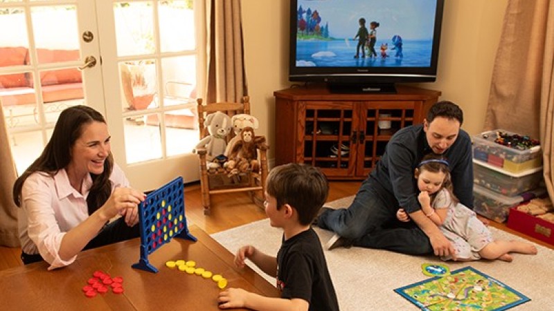 In a living room, a mother and son play Connect Four and a father and daughter play Chutes and Ladders.