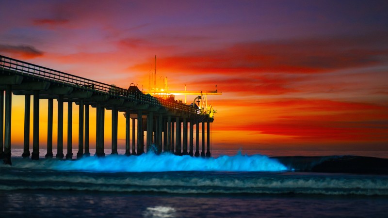 A pier enters the left side of frame. To its right, breaking waves glow neon blue. Behind it there is a red, yellow, pink and purple sunset.