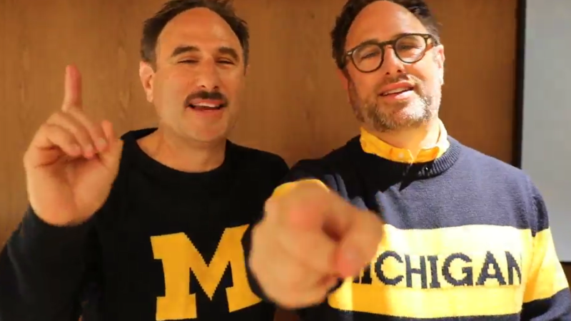 A screenshot from a video of the Sklar brothers. Both wear Michigan sweaters and point towards camera.