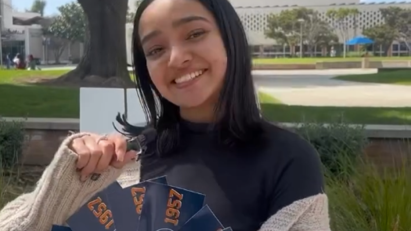 A Fullerton student holds a tiny mic in one hand and in the other holds Fullerton-branded stickers up to the camera.