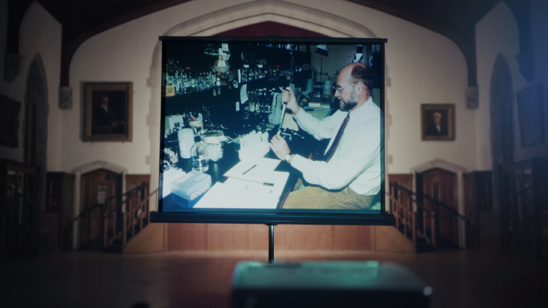In a church, on a projector screen, an old photo of a man in a lab using a pipette.