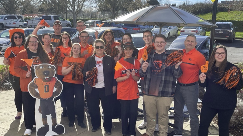 A group of people stand outside next to a parking lot wearing OSU-branded shirts and holding black- and orange-colored pom poms.