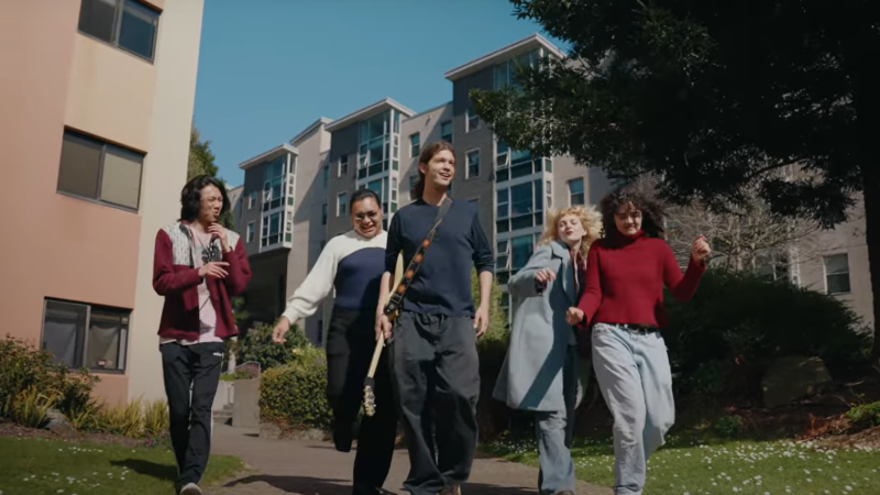 A group of five students dances down a stone pathway on SFSU's campus. The student at center has a guitar slug across their body.