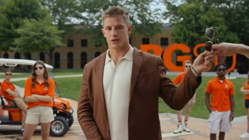 A BGSU student hands his sunglasses to someone off screen. Behind him stand other students, all wearing matching orange polos.
